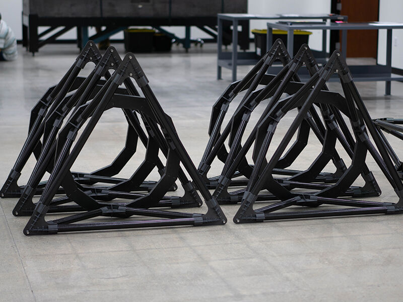 Image of TMT metrology fixtures lined up on the floor of a manufacturing area.  There are two rows of three and the viewer is above and looking down towards to the precision fixtures. The hardware appears as black circular shapes with a hollow center that are attached at three major points to black triangular shapes.